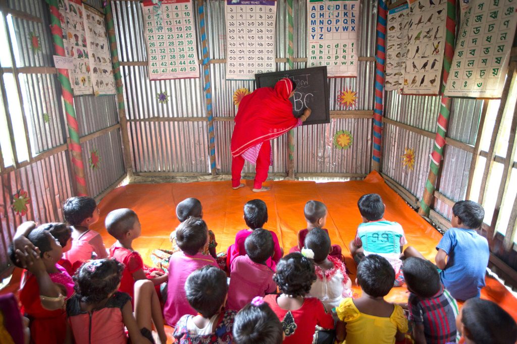 Classroom with young kids watching a female teacher write on a small chalkboard, there are alphabet, animal and bird posters attached to the walls.